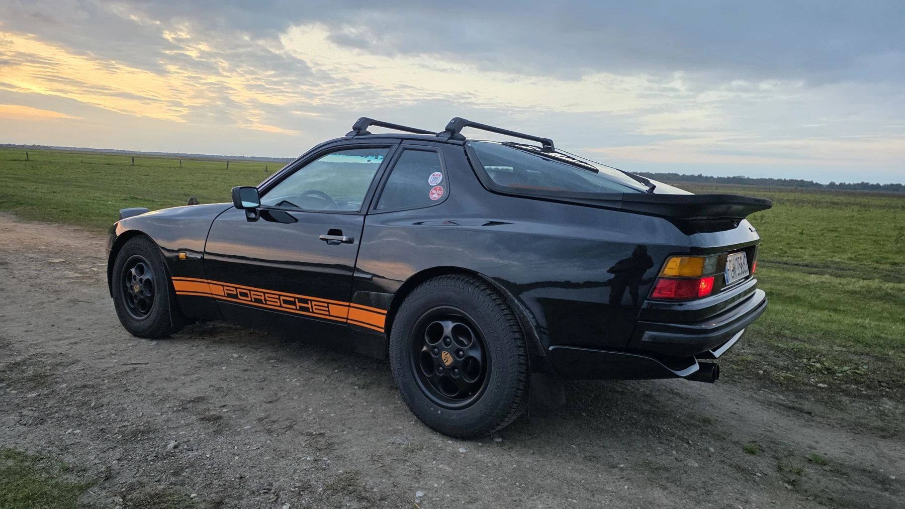 Schwarzer Porsche 944 mit orangefarbenem Porsche-Seitenstreifen und AT-Reifen auf einem Feldweg