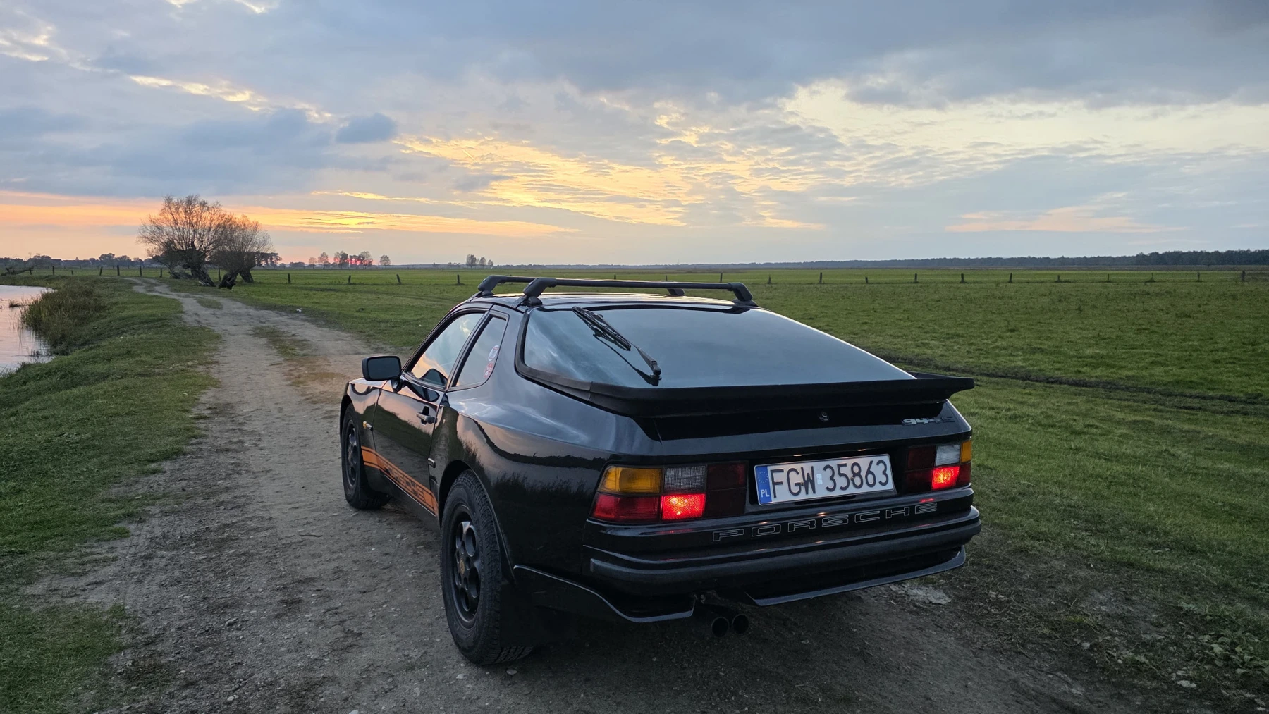 Heckansicht des schwarzen Porsche 944 auf einem Feldweg mit weitem Horizont bei Sonnenuntergang
